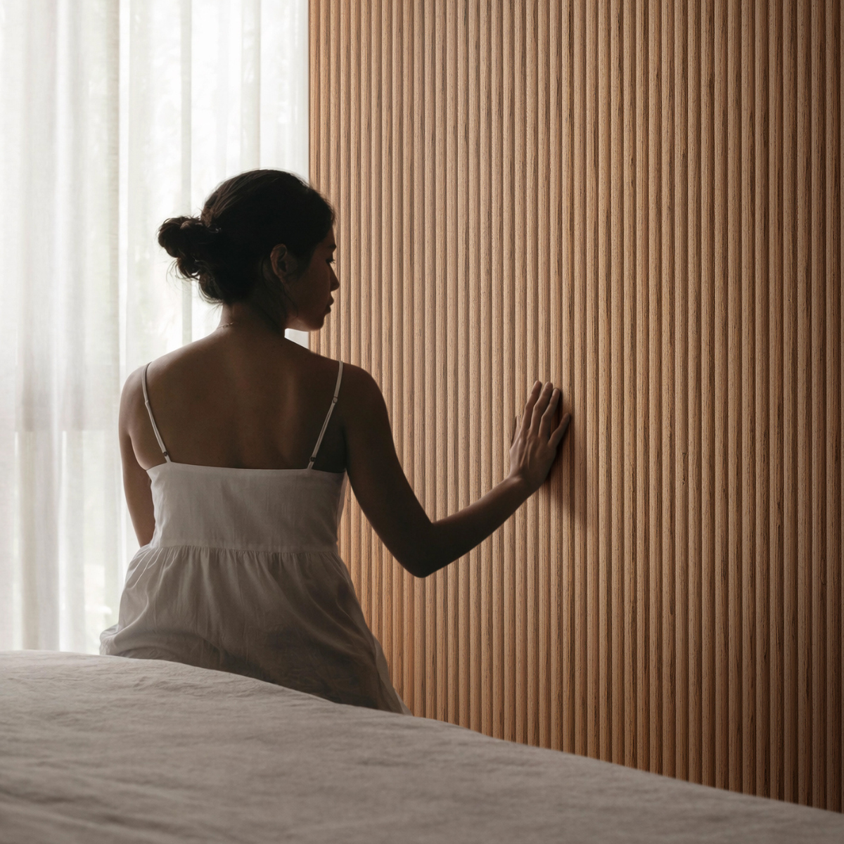 Grooved Panels της XYLOKAT ως επιφάνεια τοίχου σε υπνοδωμάτιο, με άτομο που αγγίζει την αυλακωτή ξύλινη υφή.